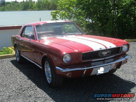 Red 1965 Ford Mustang coupe with white racing stripes, parked on gravel