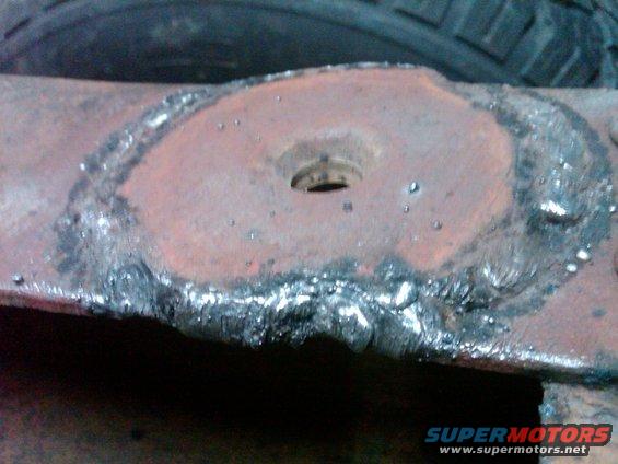 Close-up of a rusty metal part with rough welding around a central hole. A tire is visible in the background