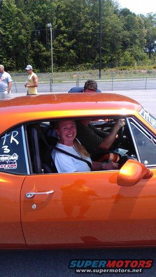 Woman in passenger seat of orange muscle car, driver visible behind wheel. Stickers on window