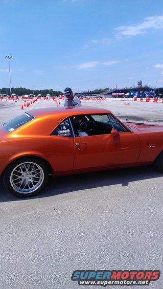 Orange 1967 coupe with aftermarket wheels and racing decals, viewed from the side at a track
