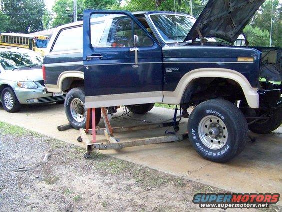 Dark blue Ford Bronco on jack stands with hood open, another vehicle parked behind it