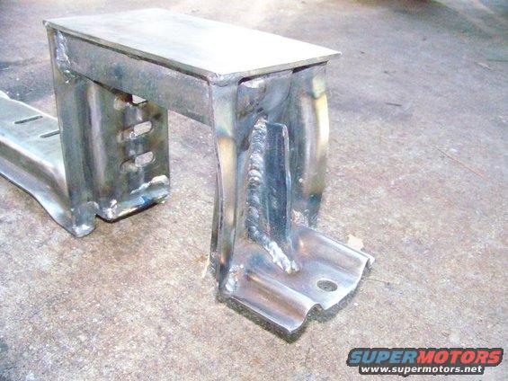 Close-up of a welded metal bracket with holes, sitting on a concrete floor. Supermotors.net logo in corner