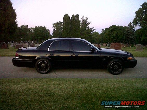 Black sedan parked on a road with trees and gravestones in the background