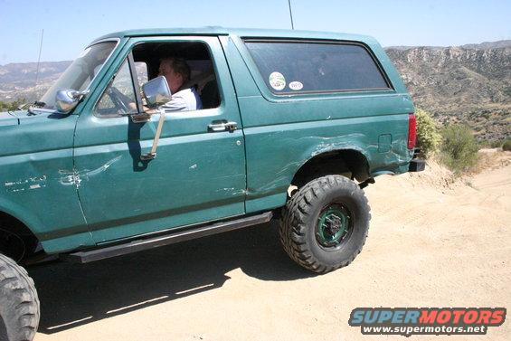Man driving green 1991 Ford Bronco on dirt road with mountains in background
