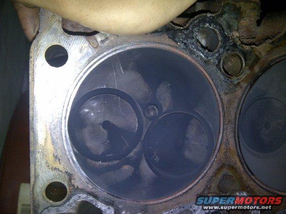 Close-up of an engine cylinder head with two valves visible inside a cylinder. Some carbon buildup is present