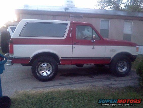 Side view of a red and white 1989 Ford Bronco parked on pavement. Spare tire mounted on the rear