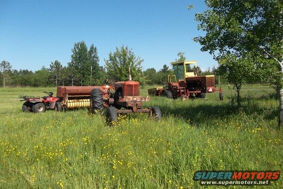Red ATV parked next to a baler and a tractor in a grassy field with a yellow combine in the background