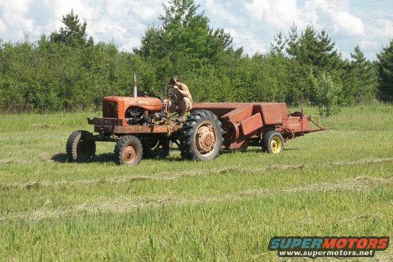 Orange tractor pulling a red baler in a grassy field. A person sits on the tractor