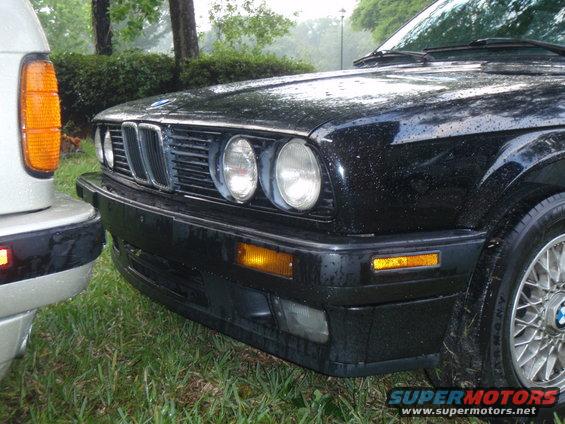 Black 1991 BMW 3 Series front bumper and grille, wet from rain, parked next to a silver SUV