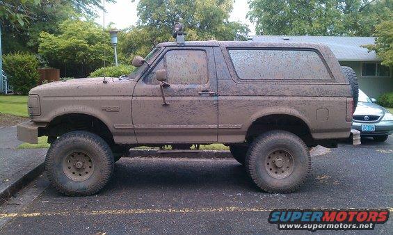 Mud-covered 1995 Ford Bronco parked on asphalt, side view. Another car is visible in the background