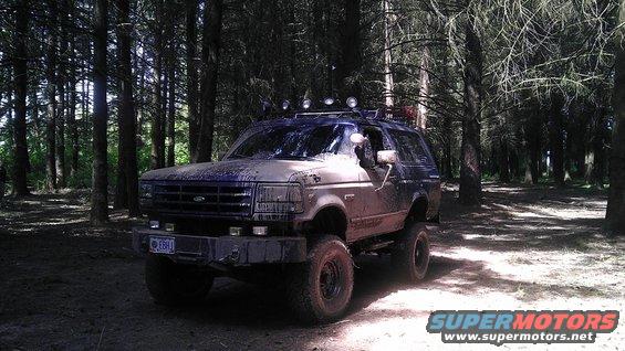 Mud-covered blue Ford Bronco with off-road tires parked in a forest