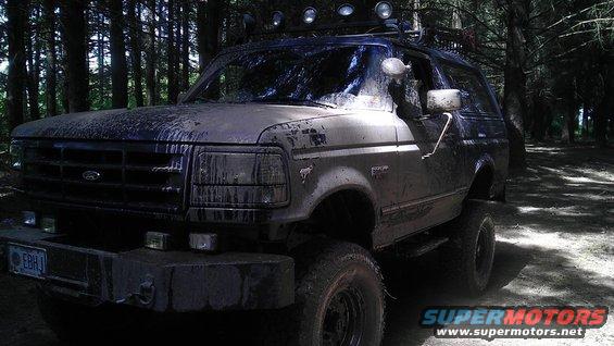 Mud-covered 1992 Ford Bronco with off-road lights on roof rack, parked in a forest