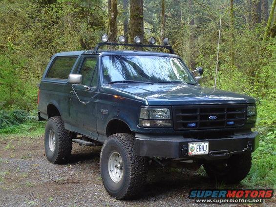 Dark blue Ford Bronco with roof lights and off-road tires parked on a dirt road in a forest