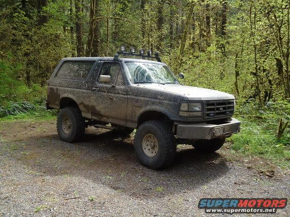 Mud-covered dark SUV with off-road tires and roof lights parked on a gravel road in a forest