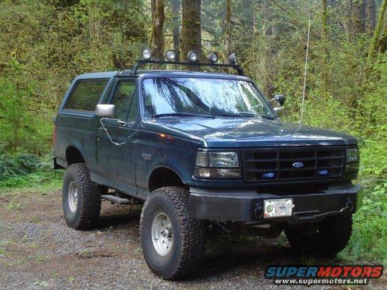 Dark blue 1995 Ford Bronco with off-road tires and roof lights parked on a dirt road in a forest