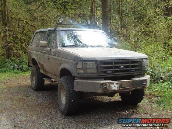 Muddy Ford Bronco parked on a dirt road in a forest. It has a light bar on the roof and a custom bumper