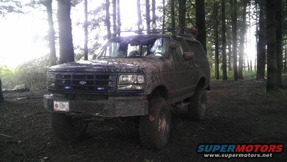 1995 Ford Bronco covered in mud, parked in a forest with trees and sunlight