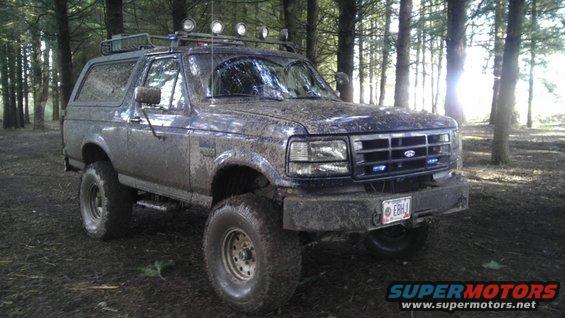 Mud-covered Ford Bronco parked in woods. Has roof lights and off-road tires