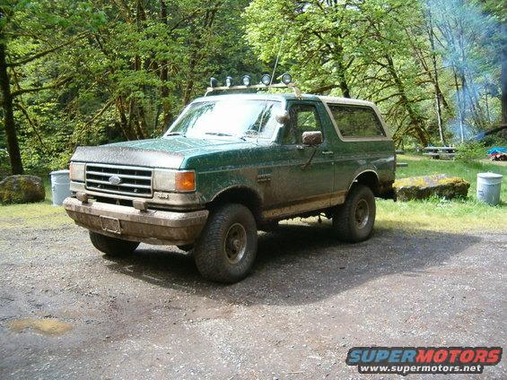 Green 1990 Ford Bronco with aftermarket lights on roof, parked on dirt road
