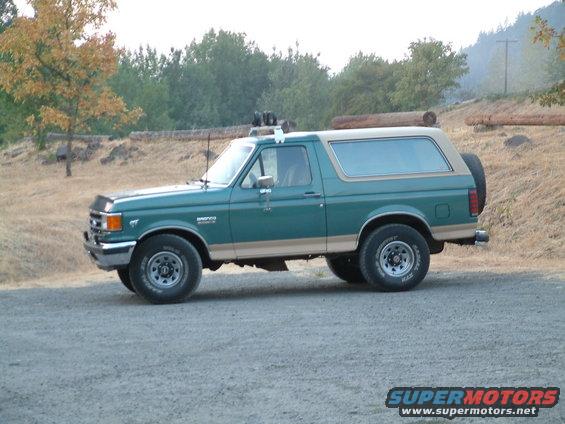 Green Ford Bronco with tan trim, parked on a gravel road with trees in the background