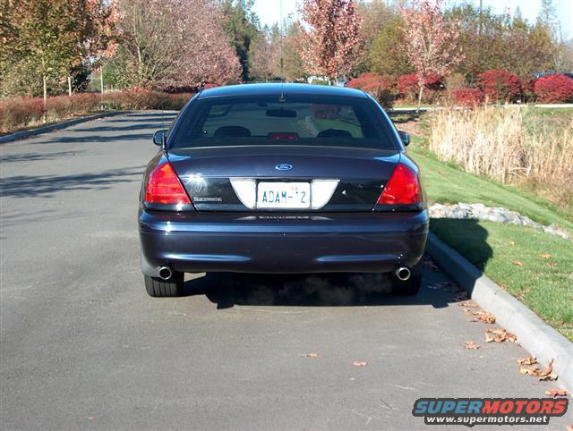 Rear view of a dark blue Ford Crown Victoria with license plate ADAM-12. The car is parked on asphalt next to a grassy embankment
