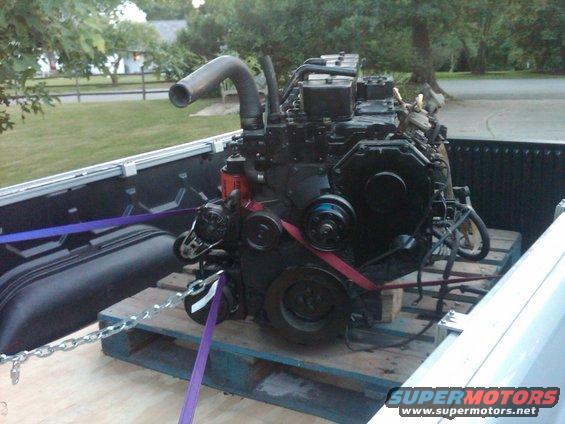 Black diesel engine on wooden pallets in truck bed, secured with purple straps and chains