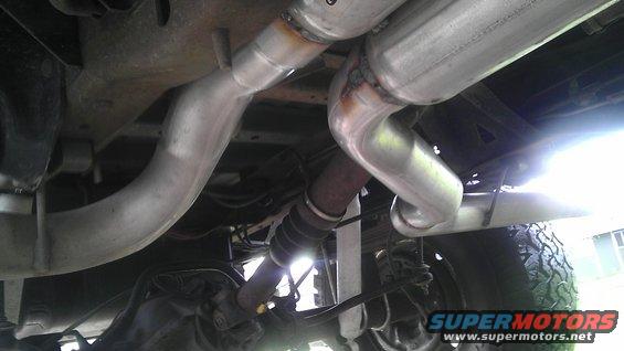 Underside view of a vehicle's exhaust pipes and muffler system. Shows a large muffler and bent pipes