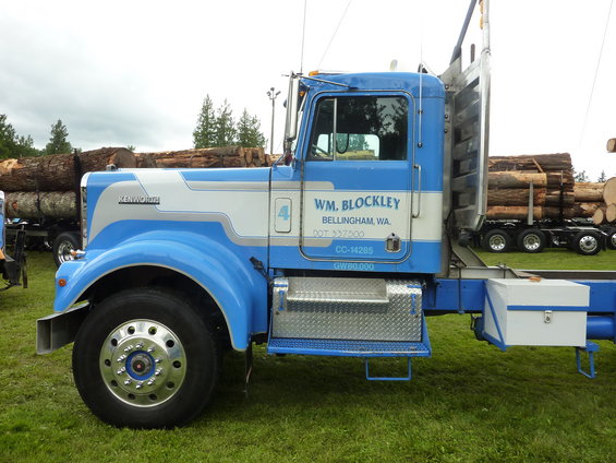 1959 Kenworth W900A logging truck, blue and white cab, carrying logs. Another log truck is visible in the background