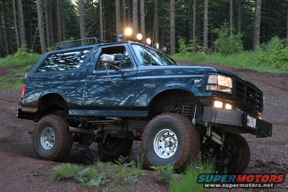 Dark teal lifted Ford Bronco with off-road tires on a dirt path in a forest