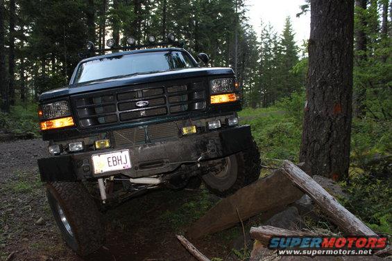 Dark green Ford truck with aftermarket lights on roof, driving over logs on a dirt trail