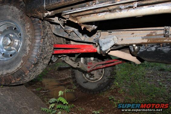 Undercarriage of a muddy off-road vehicle with red suspension components and large tires on uneven terrain