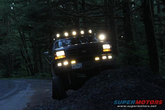 Ford truck with roof-mounted lights and aftermarket bumper lights, driving on a dirt road in a forest