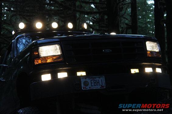 Dark Ford truck with roof lights and front bumper lights, seen at night in a forest