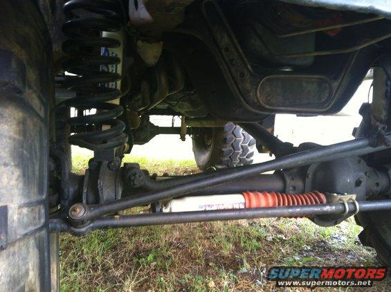 Undercarriage of a vehicle with large tires, showing suspension and steering components, with a "SUPERMOTORS" watermark
