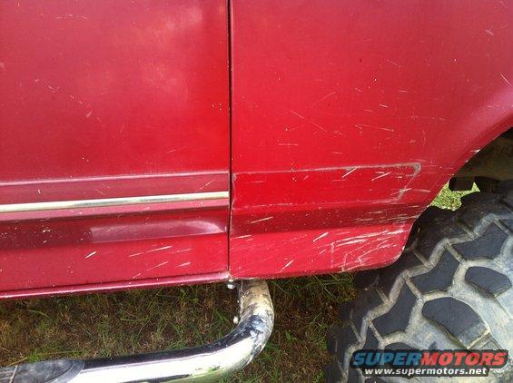 Red vehicle side panel with chrome trim, large off-road tire, and visible scratches