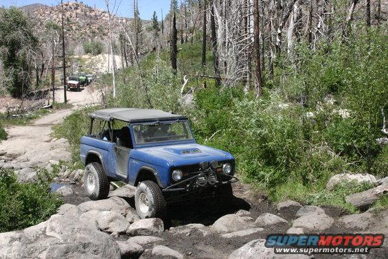 Blue first-gen Ford Bronco navigating a rocky trail with other vehicles behind it