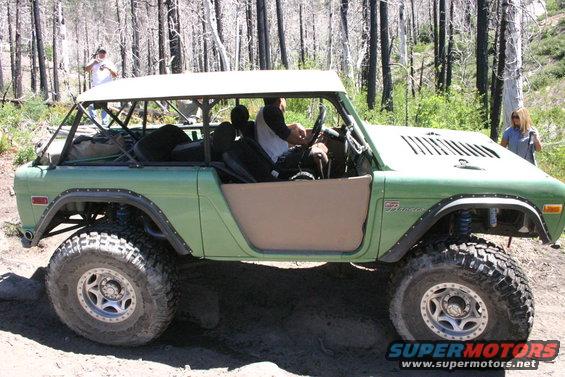 Green Ford Bronco with a white roof and tan doors, lifted with large tires, driving on a dirt trail