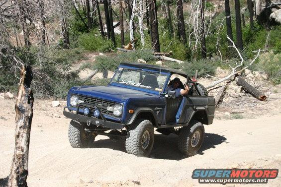 Blue first-generation Ford Bronco with aftermarket bumper and lights driving on a dirt road in a wooded area