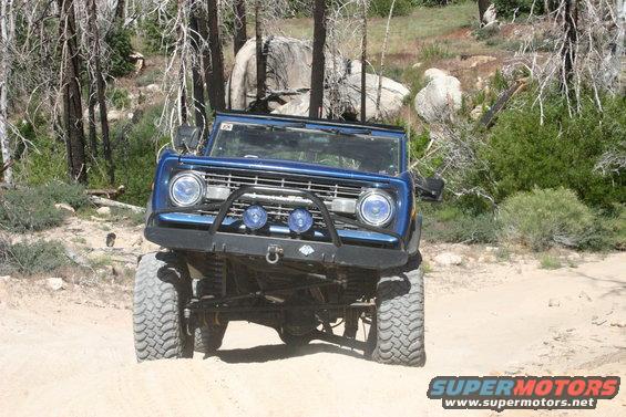 Blue Ford Bronco with off-road tires and bumper driving on a dirt road through trees