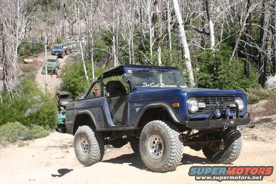 Blue Ford Bronco with off-road tires on a dirt path, front bumper with lights, "So Cal Broncos" sticker on windshield