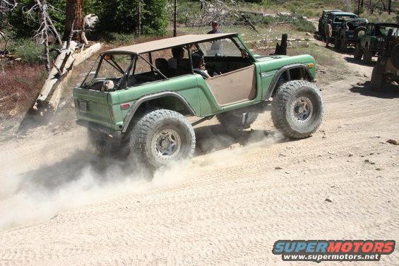 Green Ford Bronco with a tan soft top driving on a dirt road, kicking up dust