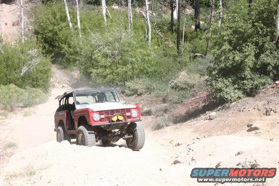 Red Ford Bronco with a white grille driving on a dirt road in a wooded area