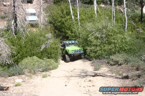 Green off-road vehicle with multiple lights on roof, driving on a dirt path surrounded by trees