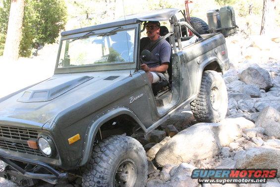 Olive green Ford Bronco with a man driving, navigating a rocky trail. It has a hood scoop and off-road tires