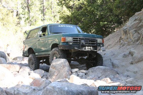 Green Ford Bronco with "ABUSED" on windshield driving over rocks on a trail