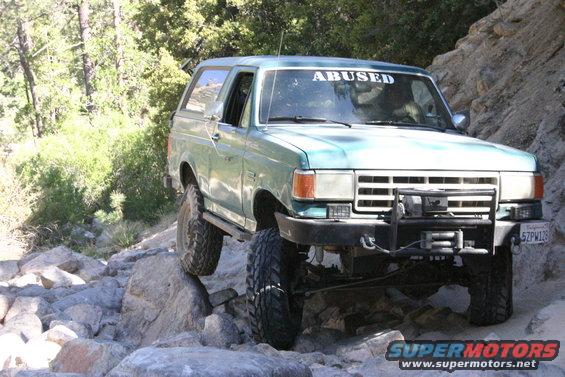 Teal Ford Bronco with "ABUSED" sticker on windshield, driving on rocks