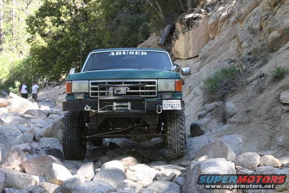 Green off-road truck with winch and "ABUSED" sticker on windshield navigating a rocky trail