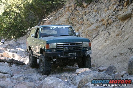 Green Ford Bronco with 'ABUSED' sticker on windshield driving over rocks on a trail