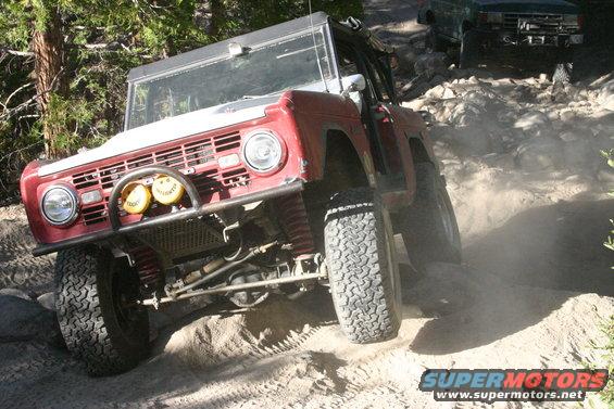 Red Ford Bronco climbing a rocky trail, kicking up dust. Another SUV is visible behind it