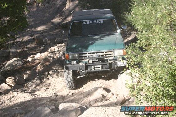 1991 Ford Bronco navigating a rocky trail, windshield banner reads 'ABUSED'. License plate 5ZPW128